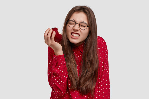 营养照片中的美女紧咬着牙齿 咬着新鲜的红苹果 穿着优雅的衬衫 戴着眼镜个人维生素新鲜