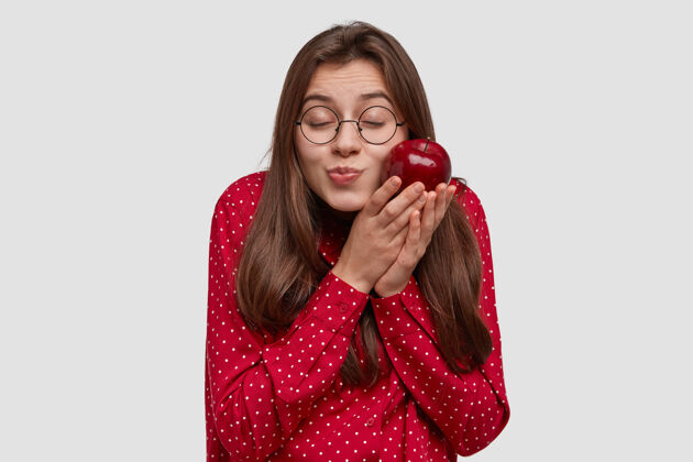 满意喜欢迷人的黑发女人 把红苹果放在脸颊边 闭上眼睛 很开心 戴着圆眼镜吃节食维生素