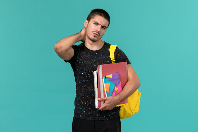 学生前视图中的男学生穿着深色t恤 背着黄色背包 手里拿着抄写本和文件 蓝色墙上有脖子疼男学生男性书籍