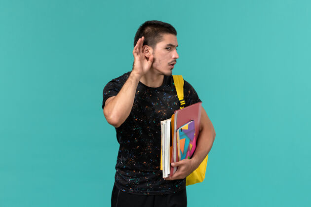学校前视图中的男学生穿着深色t恤 背着黄色背包 手里拿着抄写本和文件 试图在蓝色墙上听到声音模特听取大学