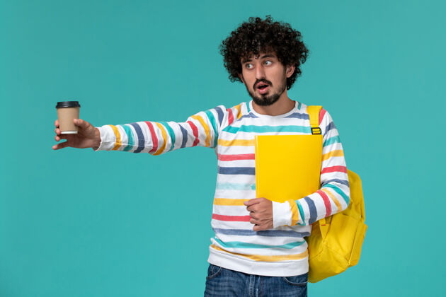 男人穿着条纹衬衫 背着黄色背包 拿着文件和咖啡的男学生在蓝色墙上的正视图男快乐咖啡