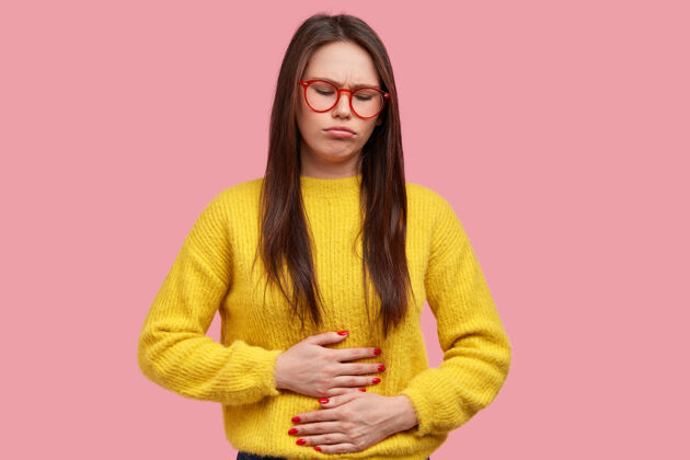 不满漂亮女人把手按在肚子上 感到疼痛和不适 有健康问题和饮食不适 穿着黄色毛衣 在粉色背景下摆姿势疾病毛衣经期