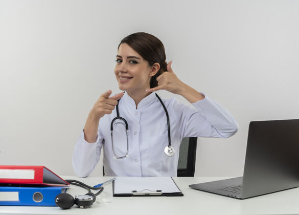 电脑微笑的年轻女医生穿着医用长袍 手持听诊器坐在办公桌旁 手持医疗工具在电脑上工作 显示呼叫电话和你的手势与复印空间坐着工具工作
