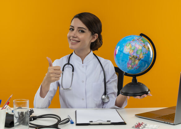 医生面带微笑的年轻女医生穿着医用长袍 手持听诊器坐在办公桌旁 手持医疗工具 在黄色背景上竖起大拇指拿着女坐着
