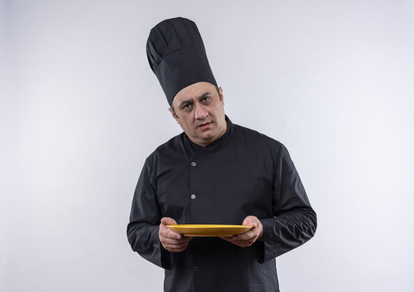 中年困惑的中年男厨师穿着厨师制服把盘子放在孤立的白墙上持有男性厨师