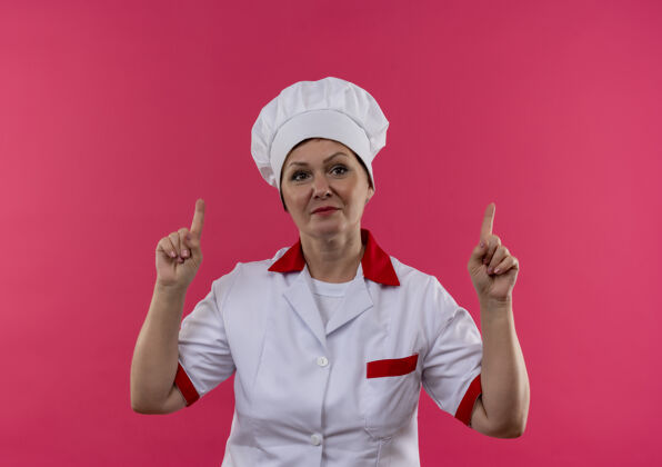 厨师身着厨师制服的中年女厨师将手指指向孤立的粉色墙壁女制服中年