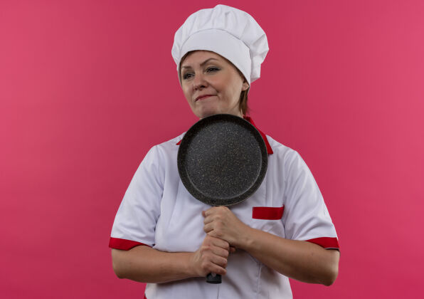 平底锅困惑的中年女厨师穿着厨师制服 拿着煎锅站在孤立的粉红色墙上厨师拿着煎着