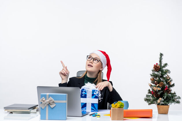 桌子圣诞气氛与戴着圣诞老人帽子和眼镜的年轻女子坐在一张桌子上拿着礼物 并显示在白色背景上面圣诞老人女人圣诞老人