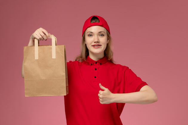 持有正面图：身穿红色制服 披风的年轻女快递员拿着快递食品包 微笑着站在粉红色的墙上包裹漂亮视图