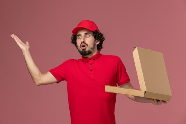 前面正面图穿着红色衬衫和斗篷的男快递员拿着空的送货食品盒 在粉红色的墙上摆姿势人人摆姿势