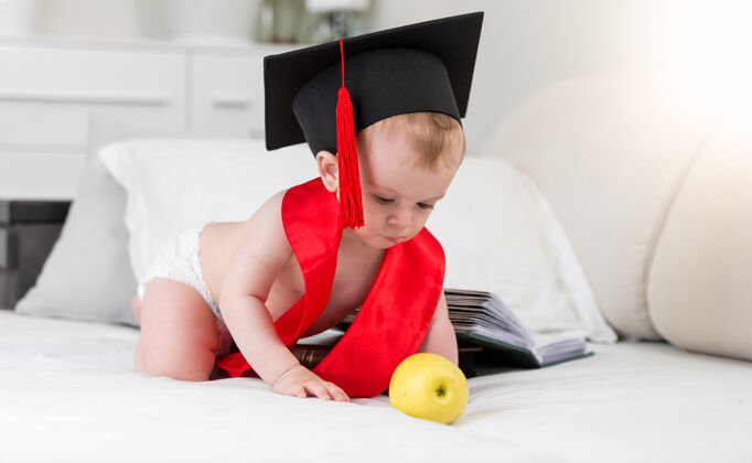 小戴着毕业帽和红丝带的神童宝宝伸手去拿苹果可爱幼儿毕业