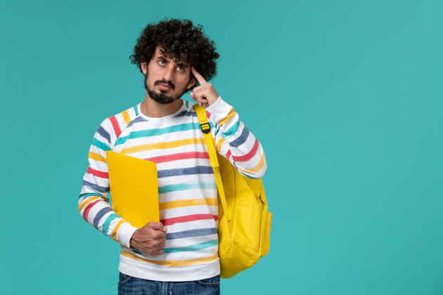 背包穿着条纹衬衫 背着黄色背包 拿着文件 在蓝色墙上思考的男学生的正视图正面大学人